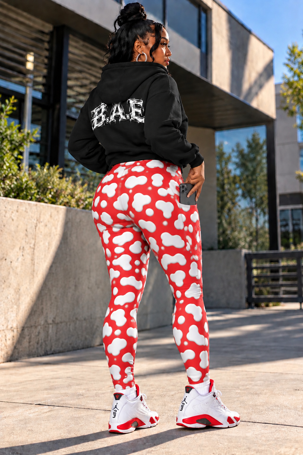 Back view of B.A.E. women's red and white cow print leggings featuring a high-waisted fit, bold printed design, stretchy fabric, and black B.A.E. hoodie, styled as a women's streetwear outfit.