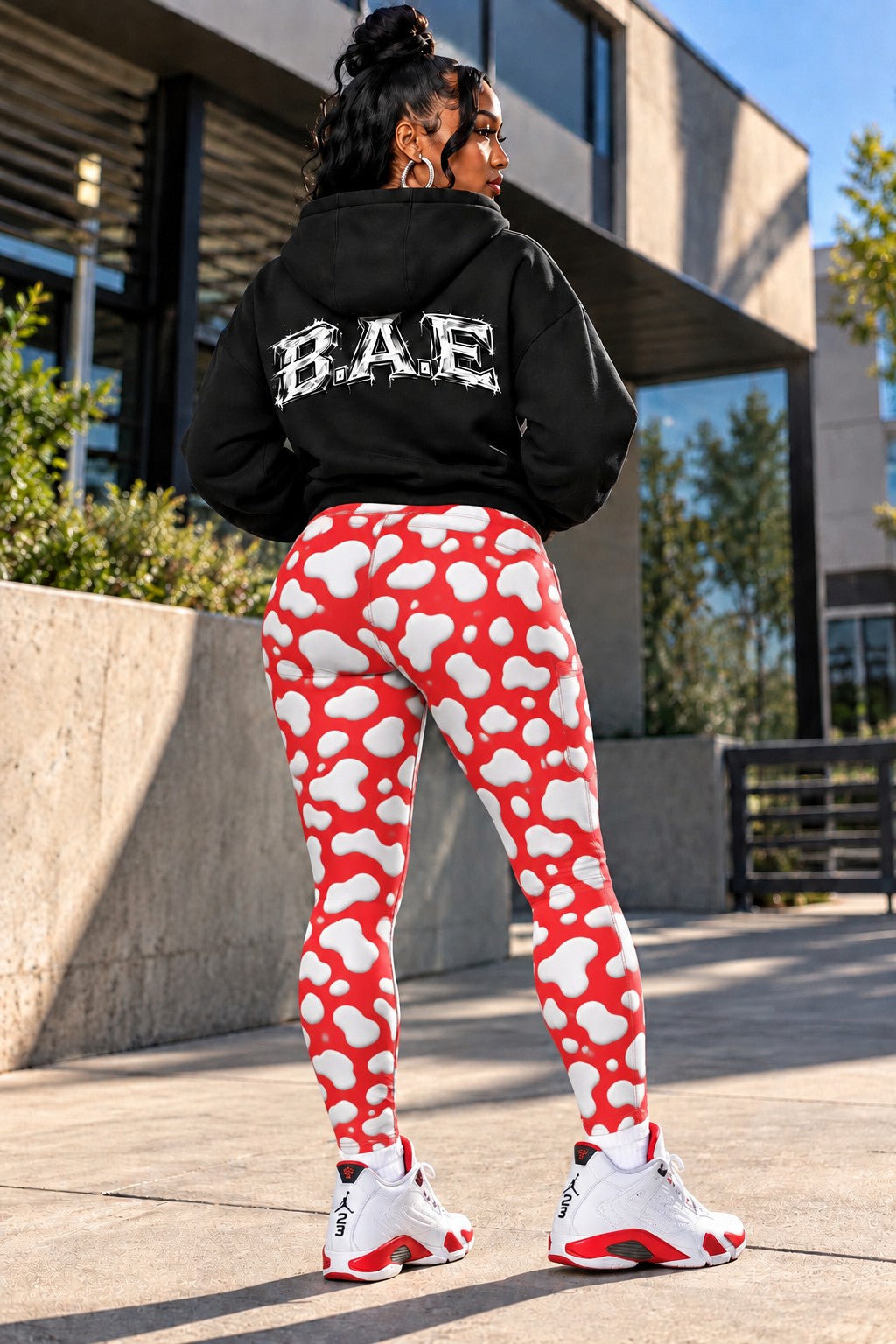 A woman standing outdoors in front of a modern building, wearing a black hoodie with 'BAE' printed in white on the back, red leggings with a white cloud pattern, and white sneakers with red accents.