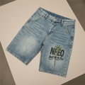 Light blue denim shorts with a distressed wash and a green and black graphic on the left leg that reads 'NFEO BUILT FOR THE FEW' with a palm tree design, shown on a plain white background.