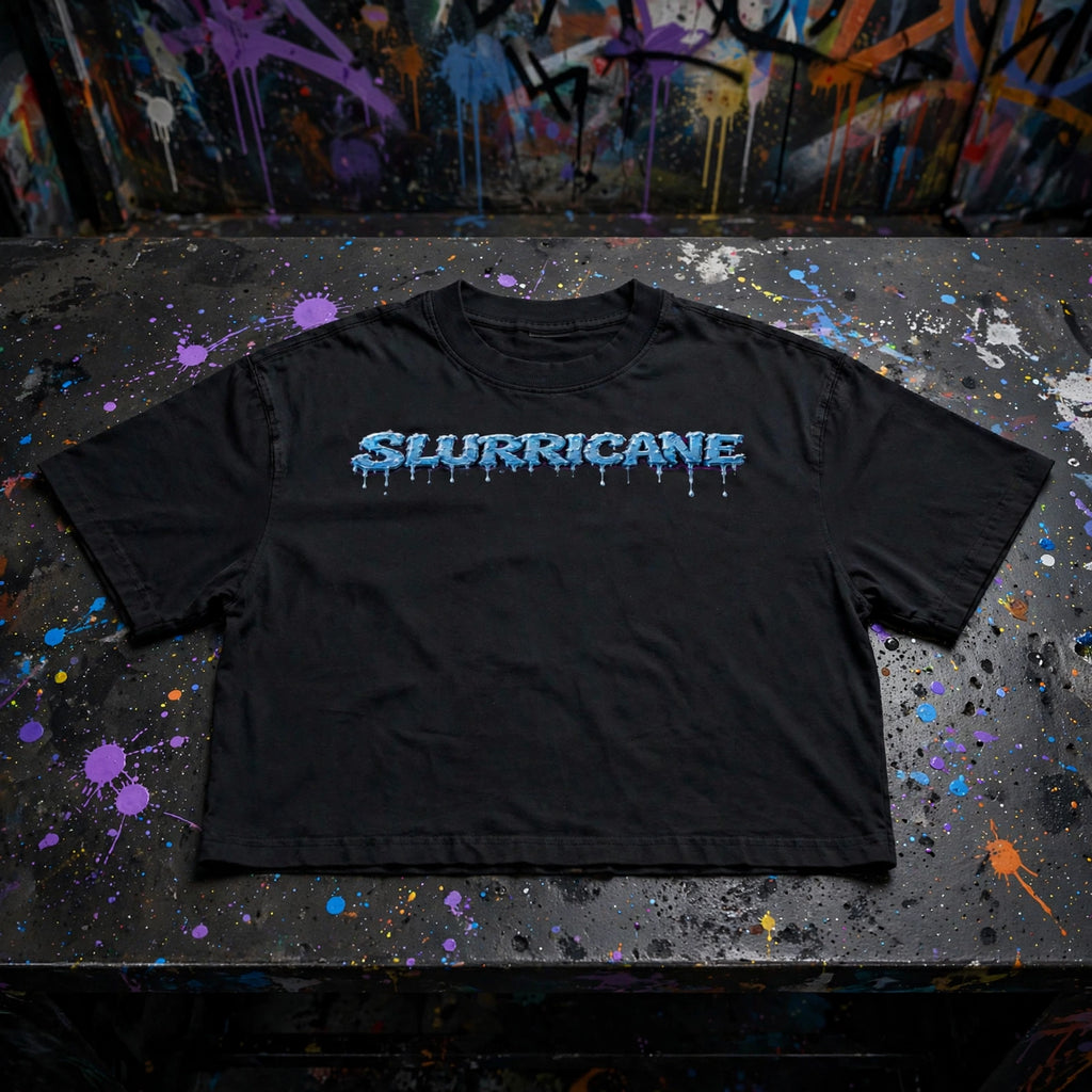 A black NFEO boxy silhouette Drop shoulders short-sleeve Tee with 'SLURRICANE' printed across the chest in a dripping blue and white font, laid flat on a paint-splattered table with a graffiti wall in the background NFEO Cannabis Strain Streetwear.