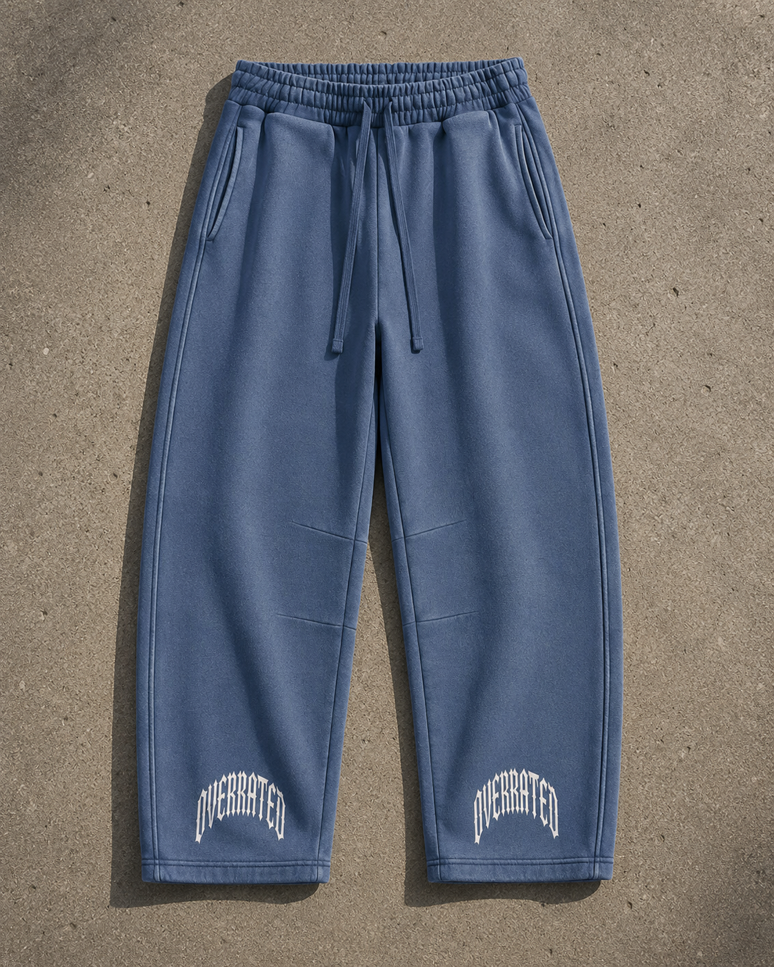 Premium blue vintage wash streetwear sweatpants flat lay featuring heavyweight 390 GSM fleece, oversized barrel-leg fit, elastic drawstring waist, side pockets, structured panel detailing, and OVERRATED typography near the cuffs. Exclusive streetwear jogger pants designed for fall and winter outfits, sneaker styling, and the NFEO OVERRATED Collection.