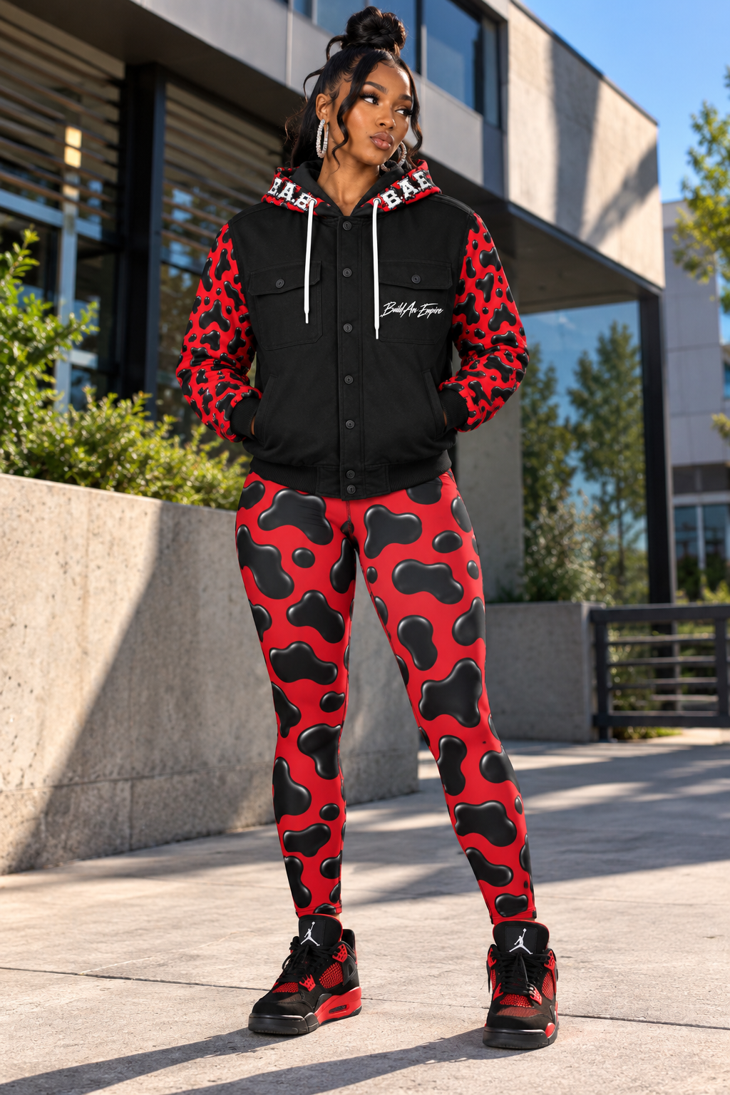 B.A.E. Build An Empire women’s red and black hooded varsity jacket styled with matching cow print leggings and black and red sneakers, showing the glossy cow print sleeves, hood artwork, front button closure, chest pockets and Build An Empire chest script.