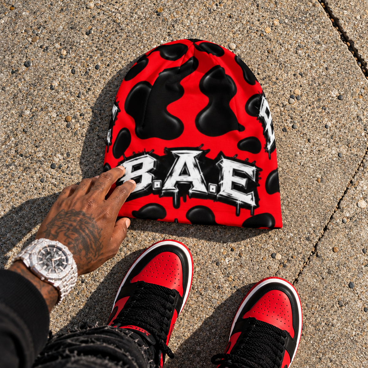 Red and black cow print B.A.E. beanie flat lay with matching red and black streetwear sneakers and male hand.