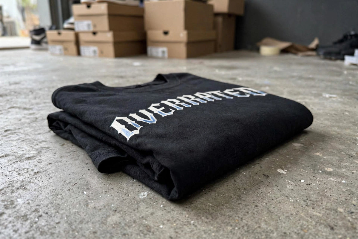 View of a folded boxy crop DropShoulders black Unisex t-shirt with a white and blue gothic-style graphic reading 'OVERRATED' lies on a concrete floor, with cardboard shoe boxes blurred in the background. NFEO Trendy Streetwear 