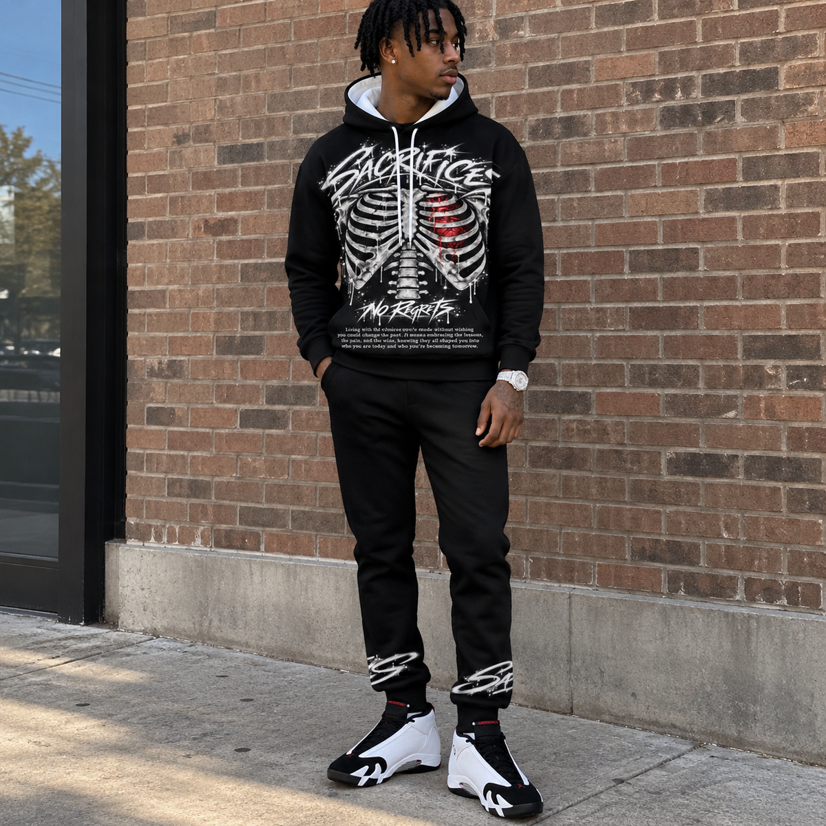 Front full-body lifestyle
Black SACRIFICES hoodie and jogger set worn by a Black male model outside a brick building, featuring a ribcage graphic with red anatomical heart, NO REGRETS design, ankle graphics, and white black red Jordan 14 sneakers.