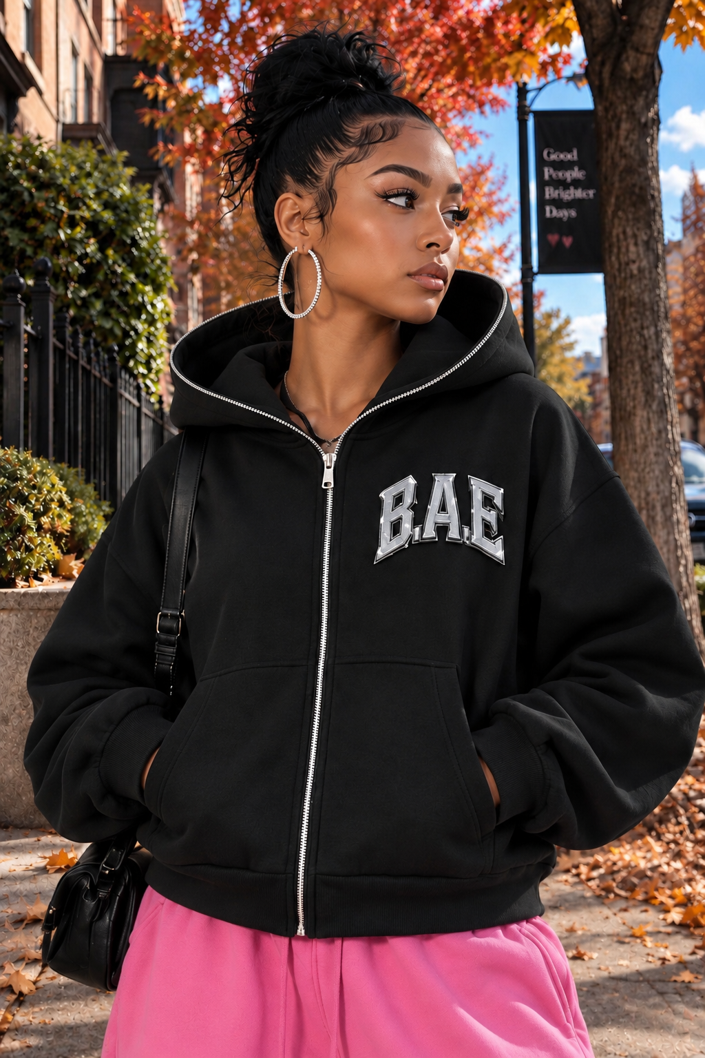 Female model wearing a black B.A.E. Built An Empire zip-up hoodie, featuring bold silver BAE chest lettering, oversized fit, full zipper, kangaroo pockets, and heavyweight fleece. Entrepreneur streetwear fashion modeled in an autumn city setting.