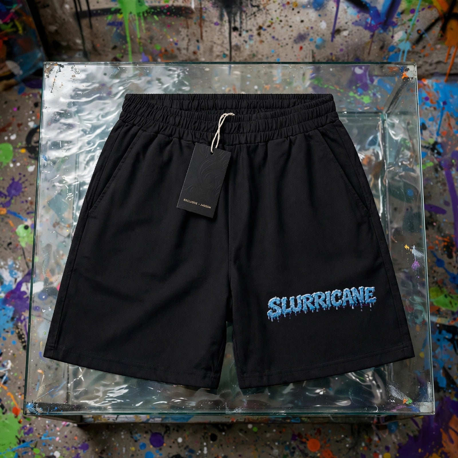 Black Slurricane boxy shorts flat lay on glass display table with hang tag - 240 GSM unisex streetwear shorts with icy blue drippy logo - ZAZA Reserve collection by Millions Pending