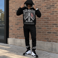Hood-up lifestyle pose
Black SACRIFICES hoodie and jogger set worn by a Black male model pulling the hood over his head outside a brick building, showing the ribcage and red heart graphic, NO REGRETS artwork, ankle graphics, and white black red Jordan 14 sneakers.