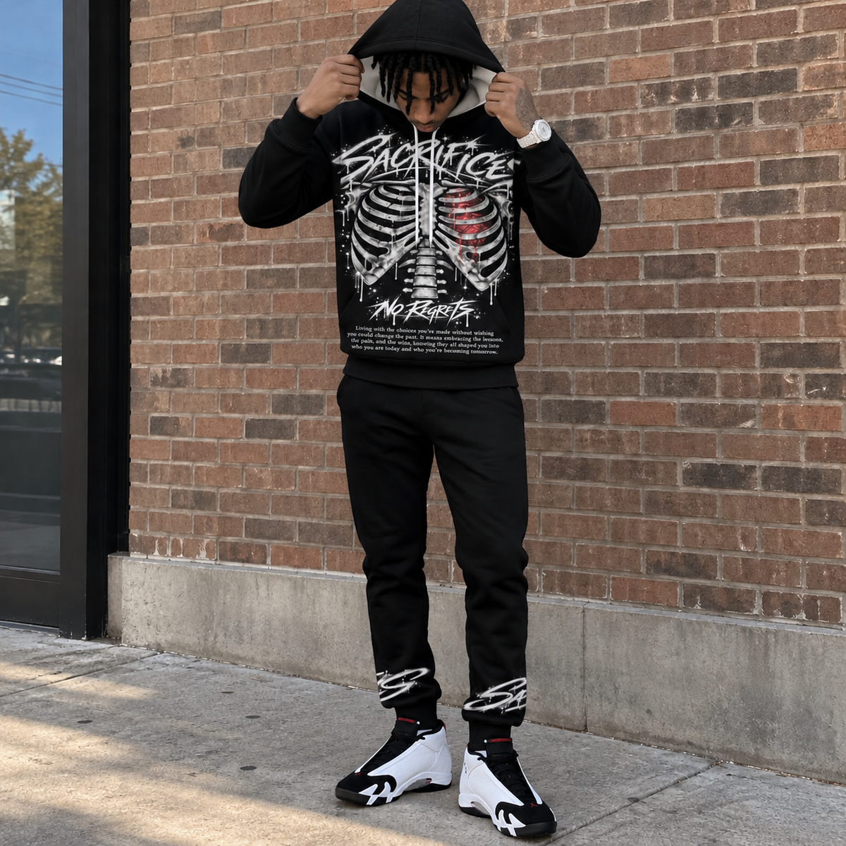 Hood-up lifestyle pose
Black SACRIFICES hoodie and jogger set worn by a Black male model pulling the hood over his head outside a brick building, showing the ribcage and red heart graphic, NO REGRETS artwork, ankle graphics, and white black red Jordan 14 sneakers.