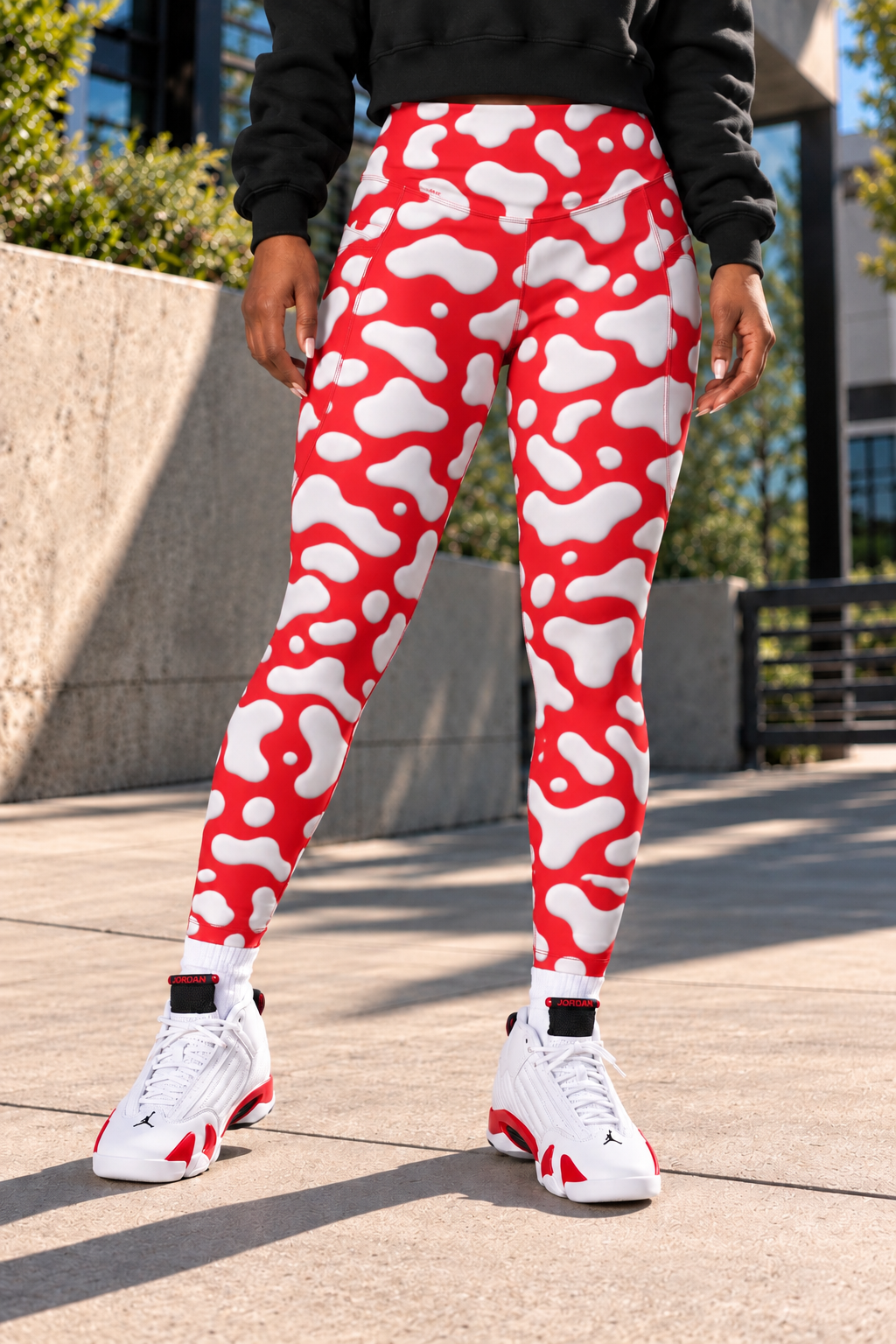 A female wearing red and white Cow patterned leggings, a black sweatshirt, and white sneakers with red accents, standing outdoors on a concrete walkway