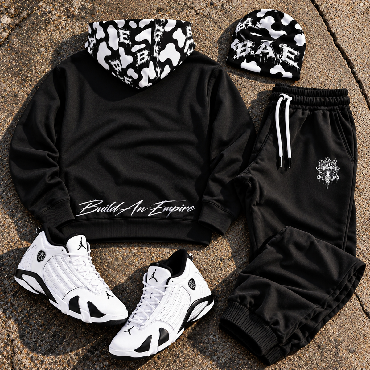 Back view of black B.A.E. streetwear hoodie and matching jogger pants featuring a black-and-white cow-print B.A.E. hood and white “Build An Empire” lower-back graphic, shown with a matching B.A.E. beanie and white sneakers.