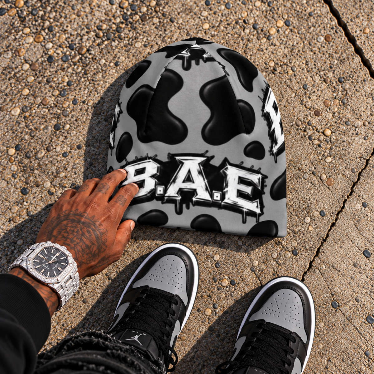 Gray and black cow print B.A.E. beanie made from stretch polyester spandex, featuring distressed Build An Empire graffiti lettering, unisex streetwear styling, and a bold all-over print design.
