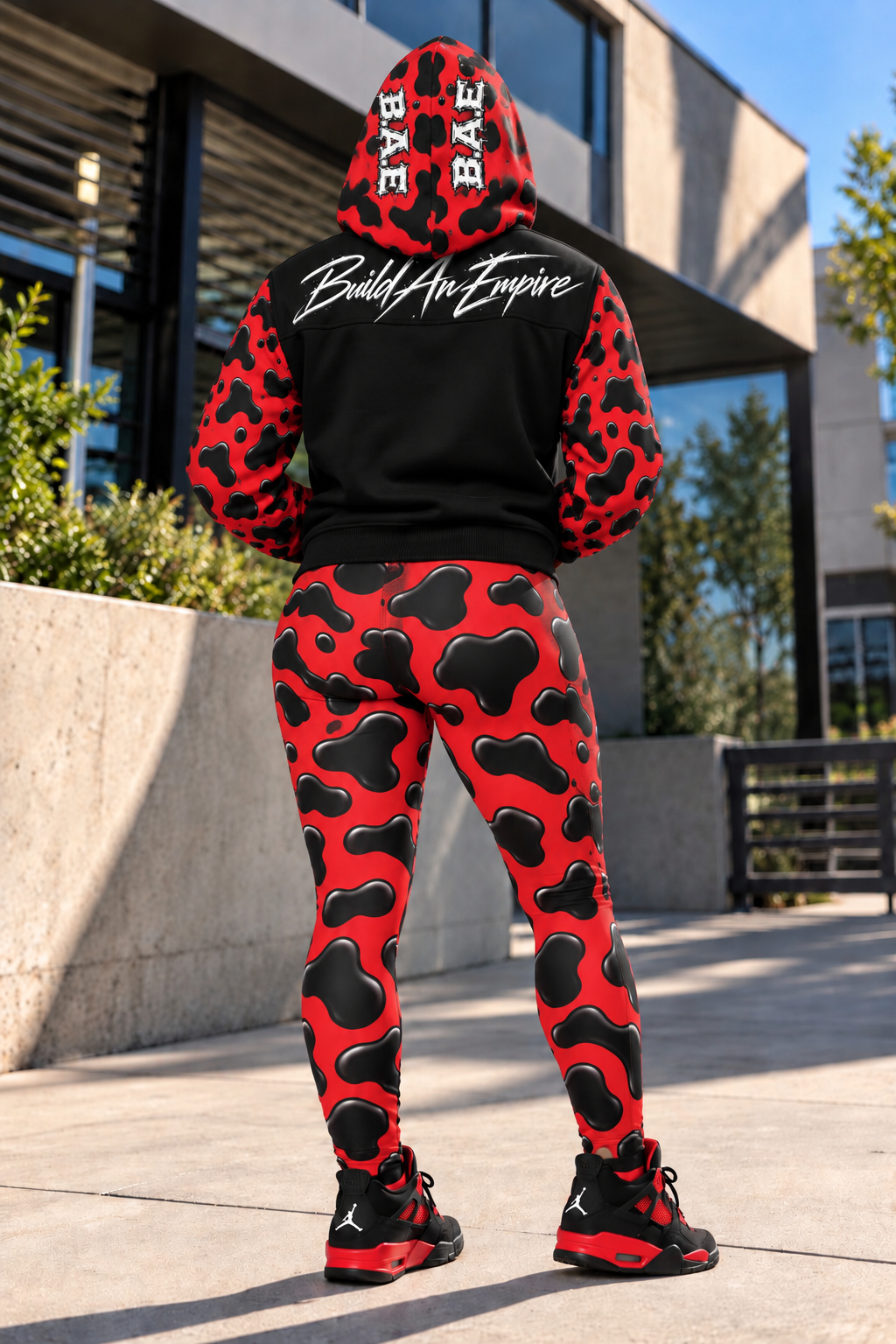 B.A.E. Build An Empire women’s red and black hooded varsity jacket modeled from the back with the hood up, glossy cow print sleeves and hood, Build An Empire script across the upper back panel, matching red and black cow print leggings and sneakers.