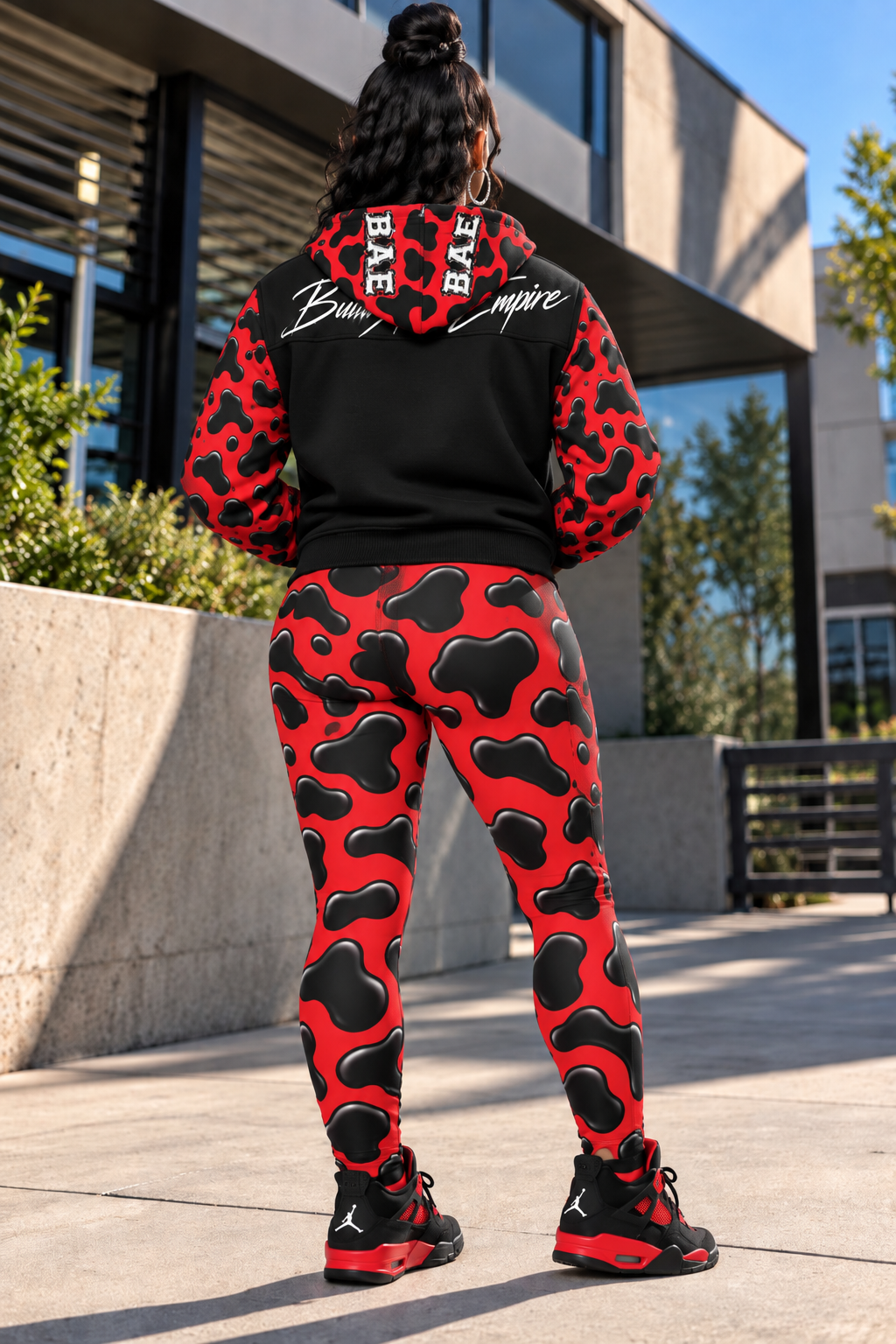 Back view female model wearing
Unisex red and black Cow pattern hooded varsity jacket shown from the back with matching cow print leggings and black and red sneakers, featuring glossy cow print sleeves and the Build An Empire script across the upper back panel.