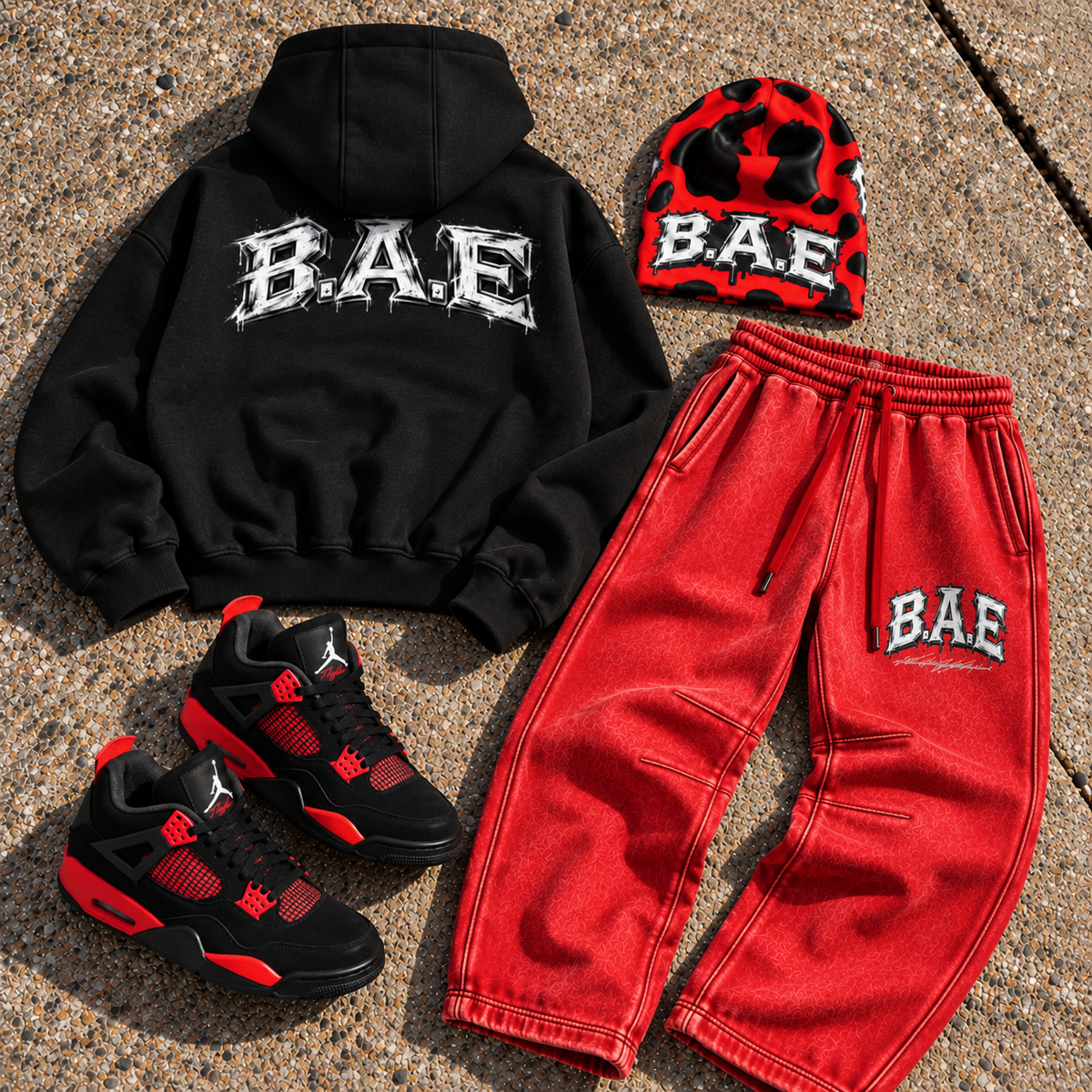 A styled flat-lay outfit set featuring a black hoodie with a white 'BAE' graphic on the back, red sweatpants with a white 'BAE' graphic on the thigh, a red and black cow printed beanie with 'BAE' text, and black and red sneakers, all arranged on a concrete surface.