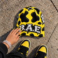 A bright yellow beanie with black cow-print spots and a black-and-white 'BAE' graphic, shown on pavement with a hand resting on it and yellow-and-black sneakers visible at the bottom.