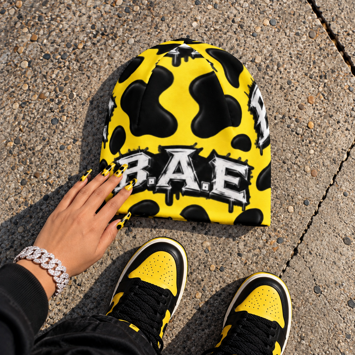 A bright yellow beanie with black cow-print spots and a black-and-white 'BAE' graphic, shown on pavement with a hand resting on it and yellow-and-black sneakers visible at the bottom.