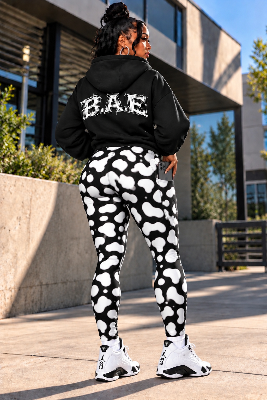 Lifestyle — Rear Side Angle
Women's black and white cow print leggings with pockets, high-waisted streetwear leggings shown from a rear side angle, highlighting the bold cow print, contoured fit and side pocket with phone, styled with a black B.A.E. hoodie and white sneakers outdoors.