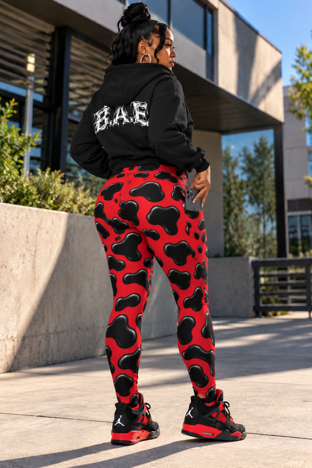 Female model turning slightly away from the camera in red and black cow print women's leggings, using the side phone pocket while wearing a black B.A.E. hoodie and black-and-red sneakers against a modern outdoor urban backdrop, highlighting the leggings' high-waisted fit and pocket placement.