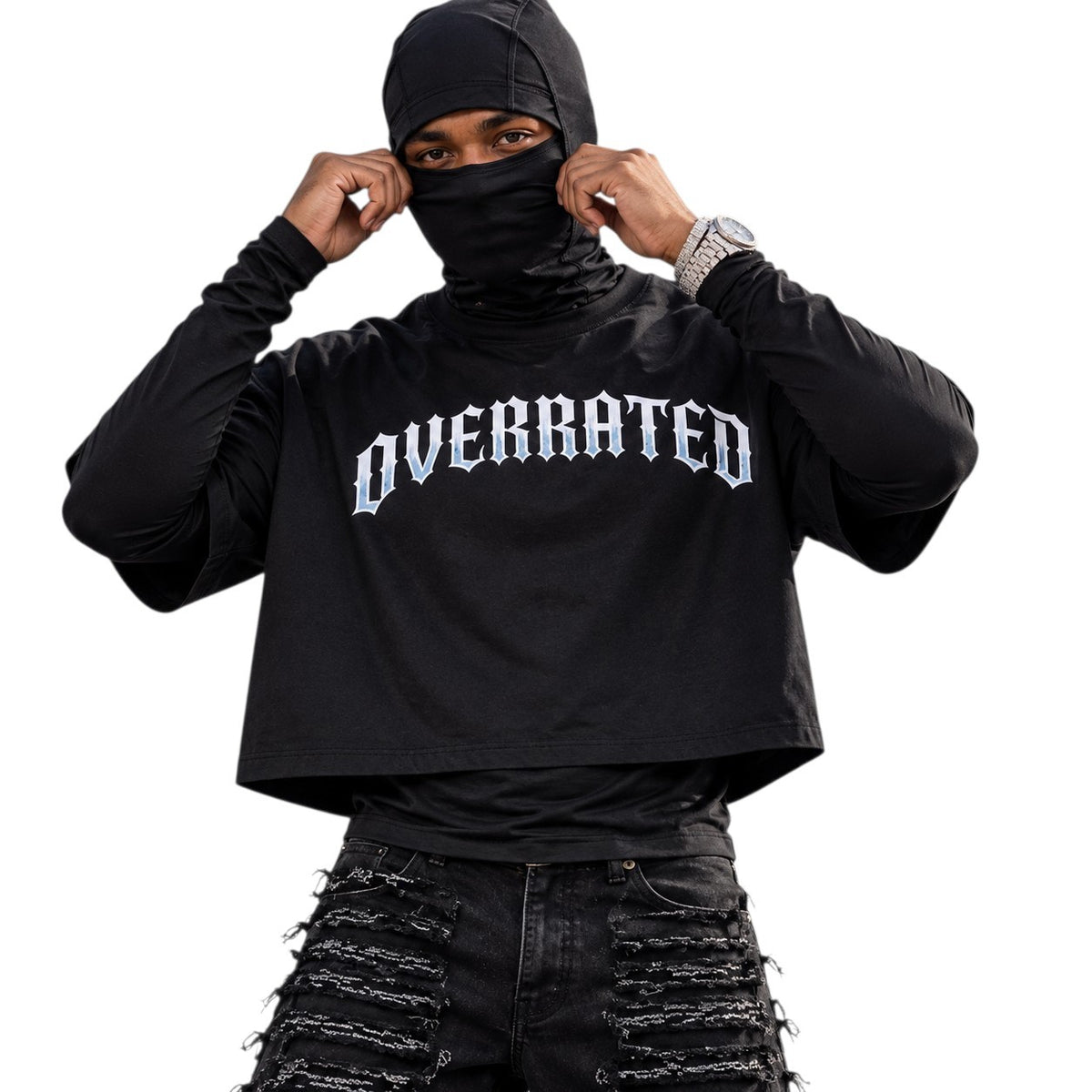 Front view:
OVERRATED black boxy crop T-shirt front view on a male model wearing a black face covering, long-sleeve base layer and heavily distressed black jeans, featuring the bold white OVERRATED chest graphic.