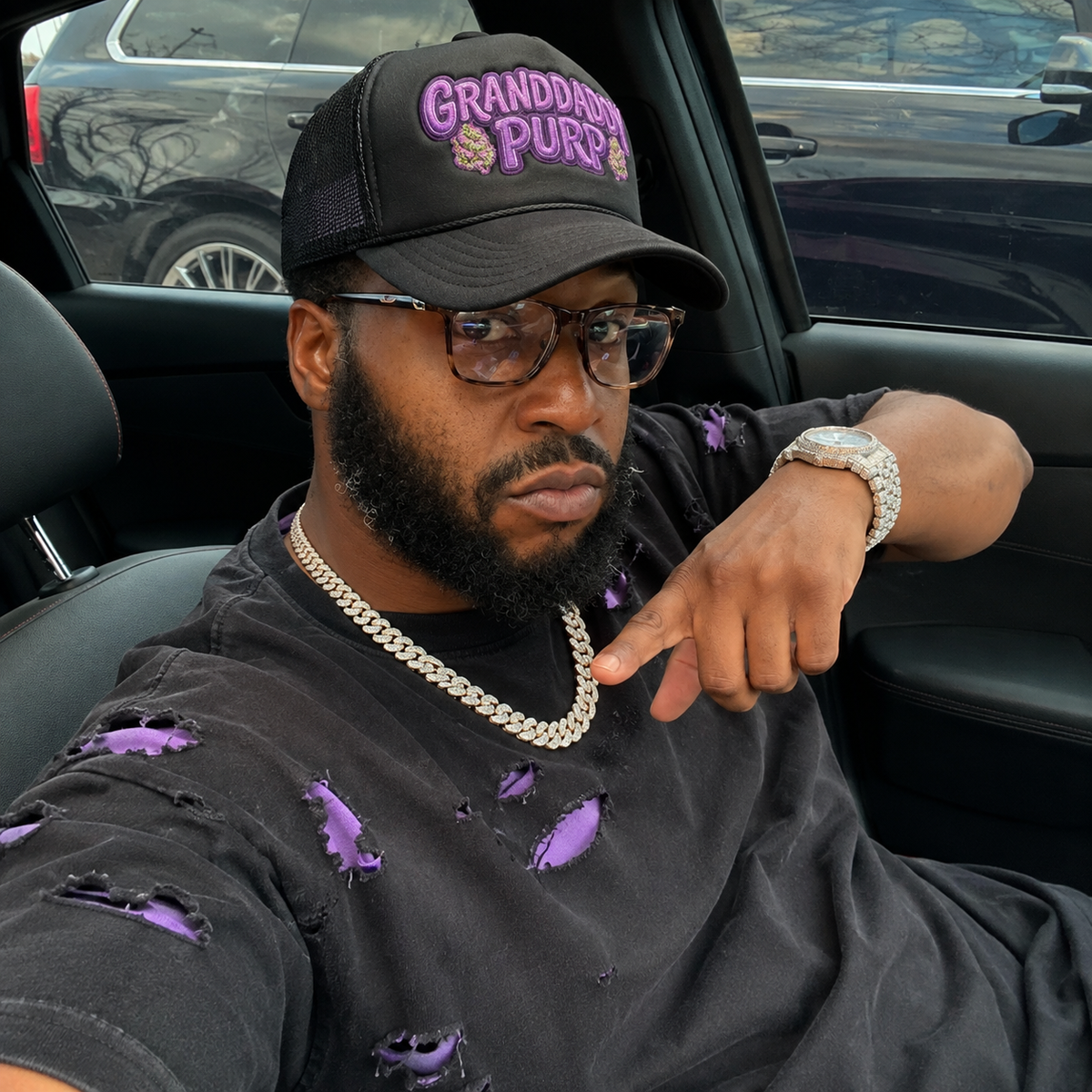 NFEO CEO wearing Our black trucker hat with purple 'Granddaddy Purp' embroidery, Cannabis Strain Hat a matching black distressed t-shirt, and a silver chain necklace, sitting inside a car.