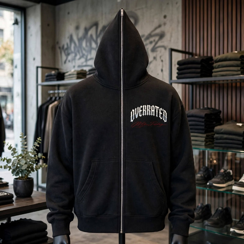 A Trendy black full zip-up hoodie displayed on a mannequin inside a retail store. The hoodie features a white arched 'OVERRATED' text graphic and a small red Millions Pending script accent on the left chest