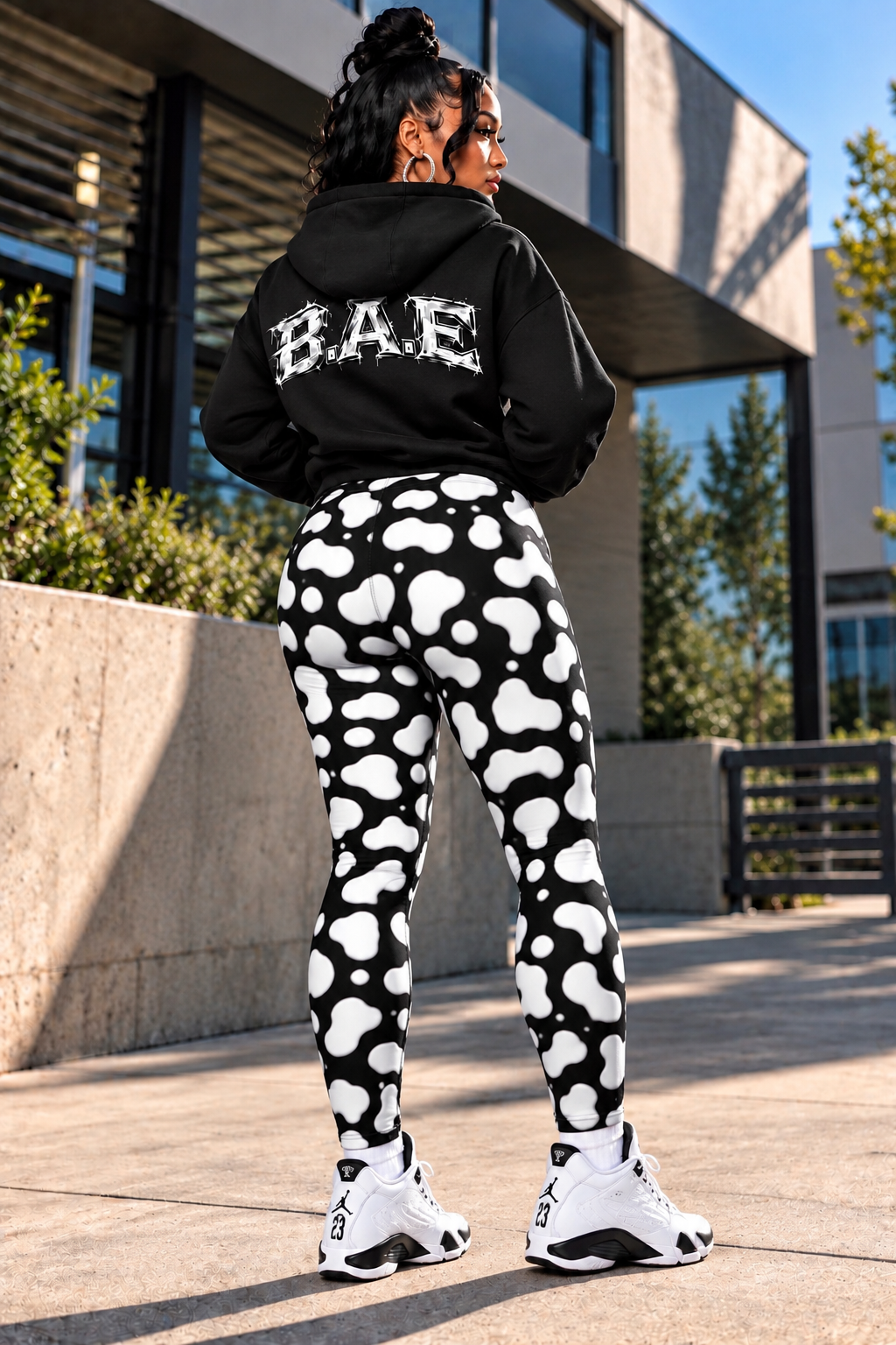 Lifestyle — Full Back Angle
B.A.E. women's black and white cow print leggings with pockets modeled from the back, showing the high-waisted fit, full-length printed leggings, side phone pocket and black B.A.E. hoodie with white sneakers in an urban outdoor setting.