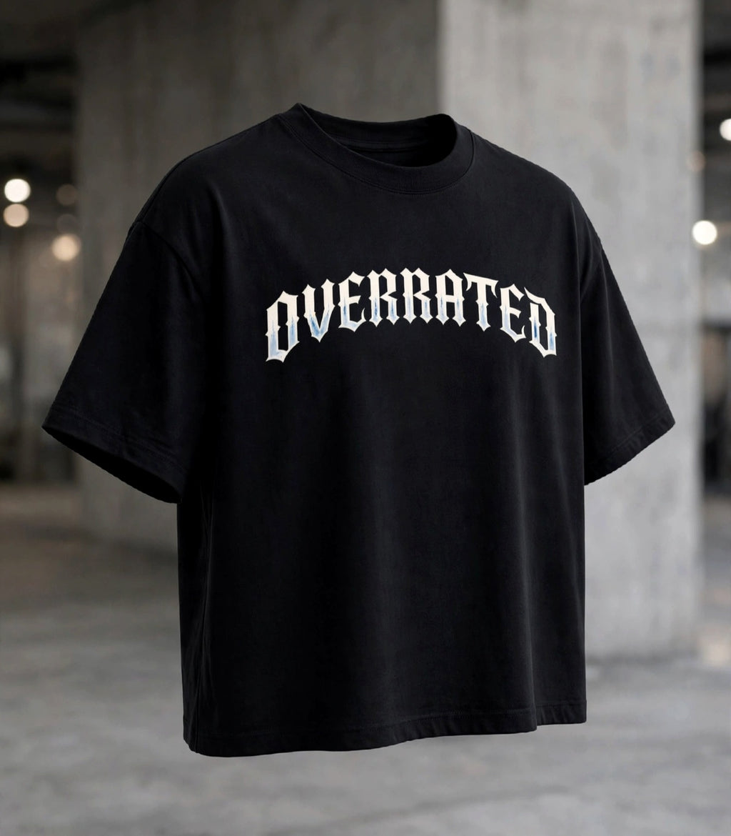 Front view of : A black Boxy Crop DropShoulders Tee oversized short-sleeve t-shirt with the word 'OVERRATED' DTG printed bold across the chest in a white gothic font, shown against a blurred concrete warehouse background. NFEO Trendy Streetwear 