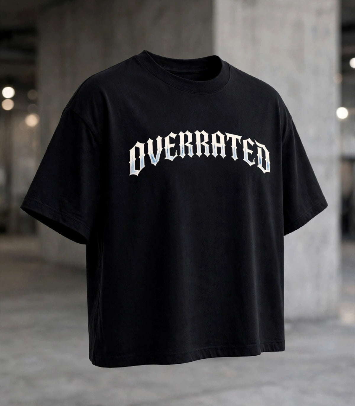 Front view of : A black Boxy Crop DropShoulders Tee oversized short-sleeve t-shirt with the word 'OVERRATED' DTG printed bold across the chest in a white gothic font, shown against a blurred concrete warehouse background. NFEO Trendy Streetwear 