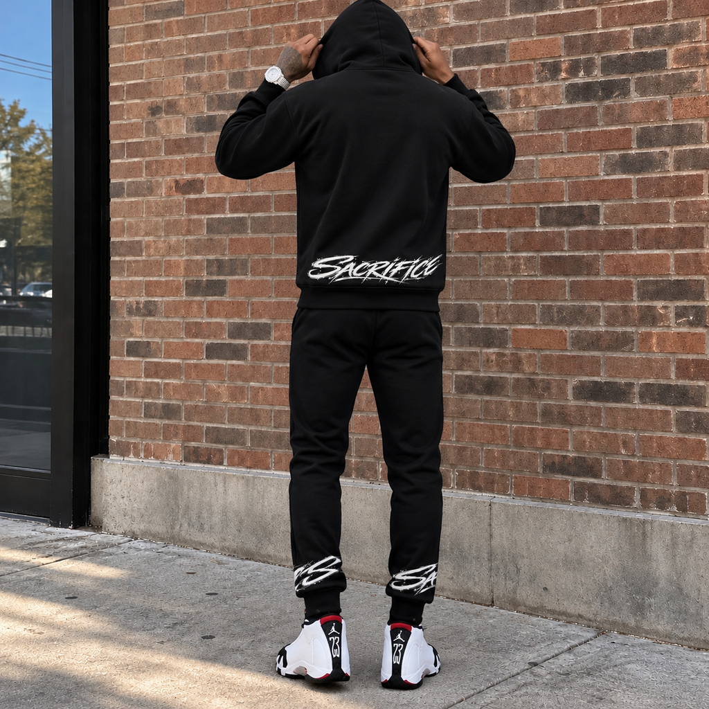 Back-facing lifestyle
Back view of a Black male model wearing the SACRIFICES black hoodie and jogger set with the thin SACRIFICES lower-back graphic, hood up, matching ankle graphics and white black red Jordan 14 sneakers outside a brick building.
