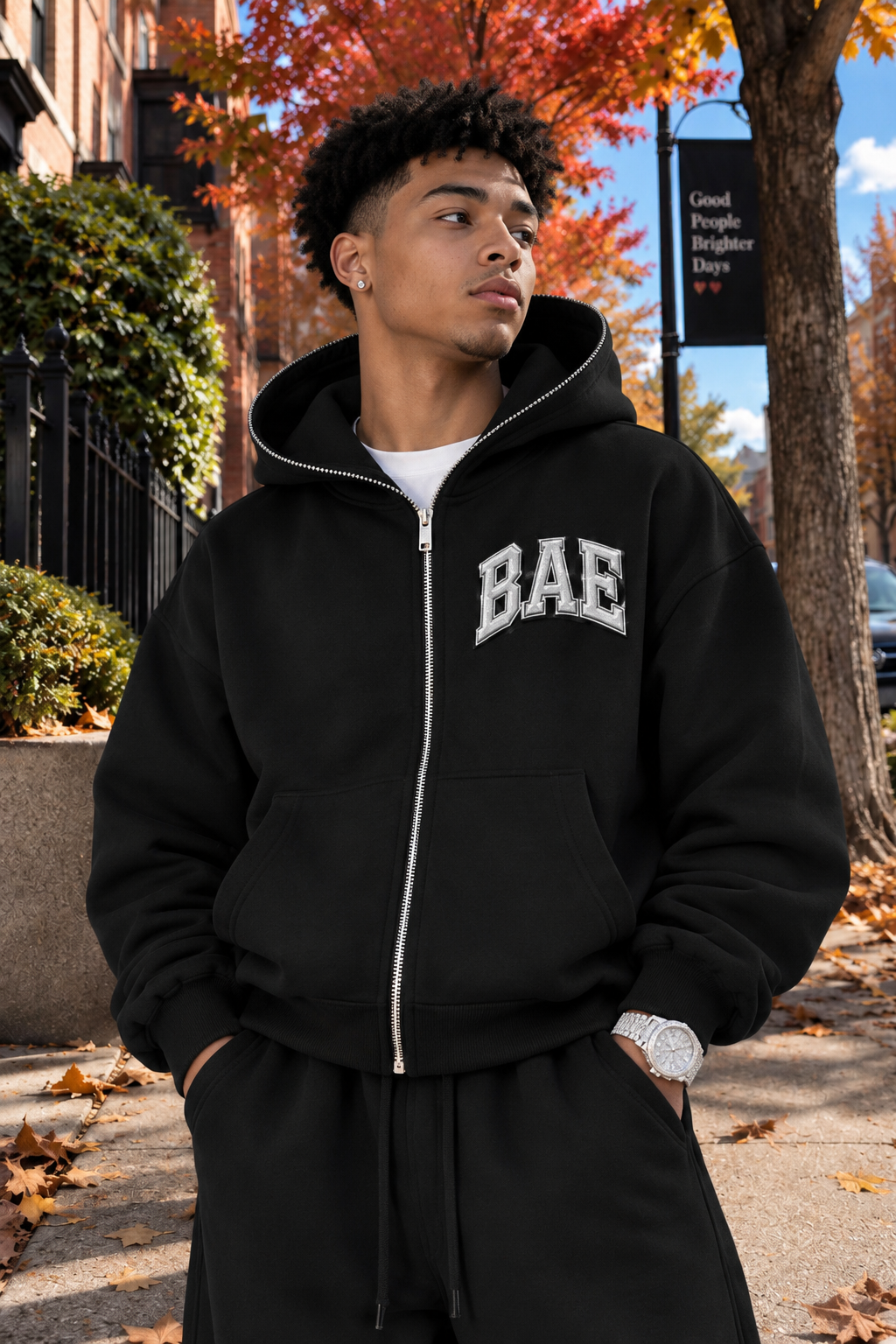 Male model wearing a black B.A.E. Built An Empire zip-up hoodie, featuring bold silver BAE chest lettering, oversized fit, full zipper, kangaroo pockets, and heavyweight fleece. Entrepreneur streetwear fashion modeled in an autumn city setting.