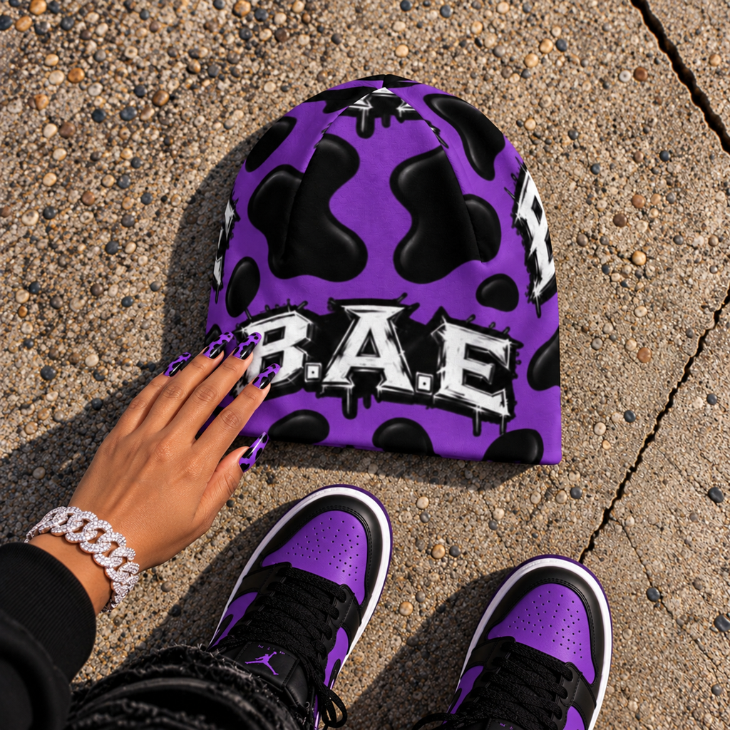 Purple and black cow print B.A.E. beanie made from stretch polyester spandex, featuring distressed Build An Empire graffiti lettering, unisex streetwear styling, and a bold all-over print design.