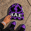 Purple and black cow print B.A.E. beanie made from stretch polyester spandex, featuring distressed Build An Empire graffiti lettering, unisex streetwear styling, and a bold all-over print design.