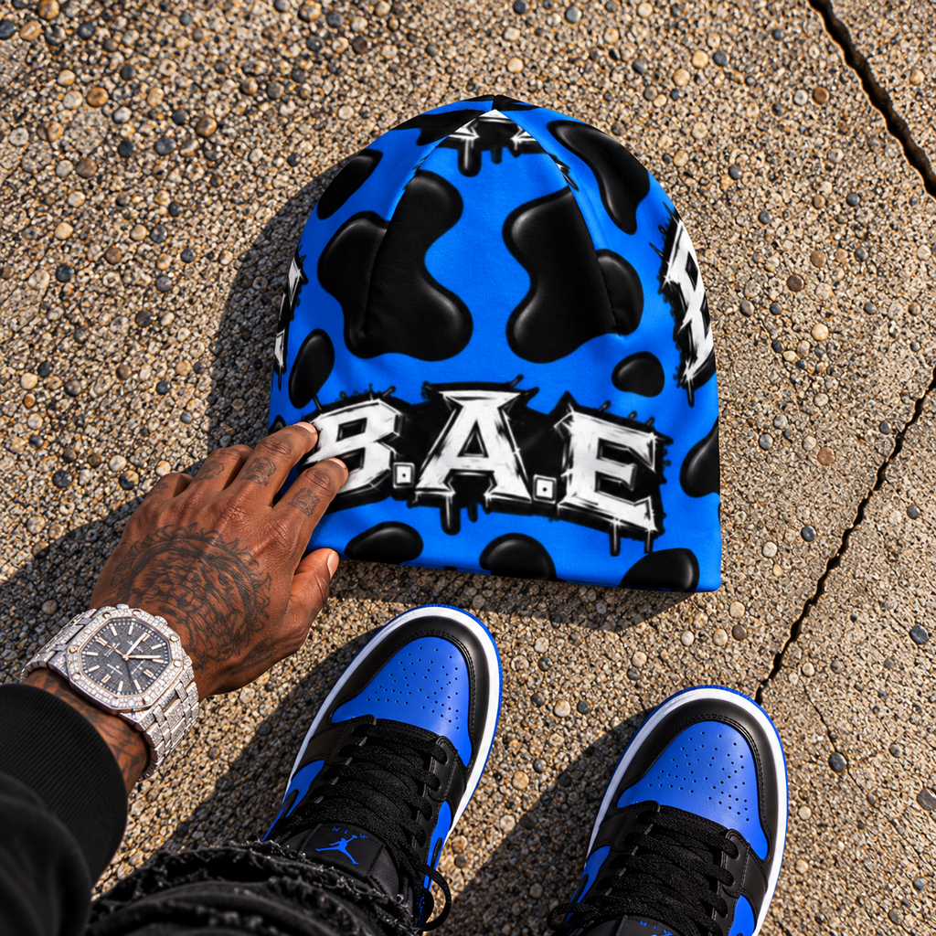 Royal blue and black cow print B.A.E. beanie made from stretch polyester spandex, featuring distressed Build An Empire graffiti lettering, unisex streetwear styling, and a bold all-over print design.