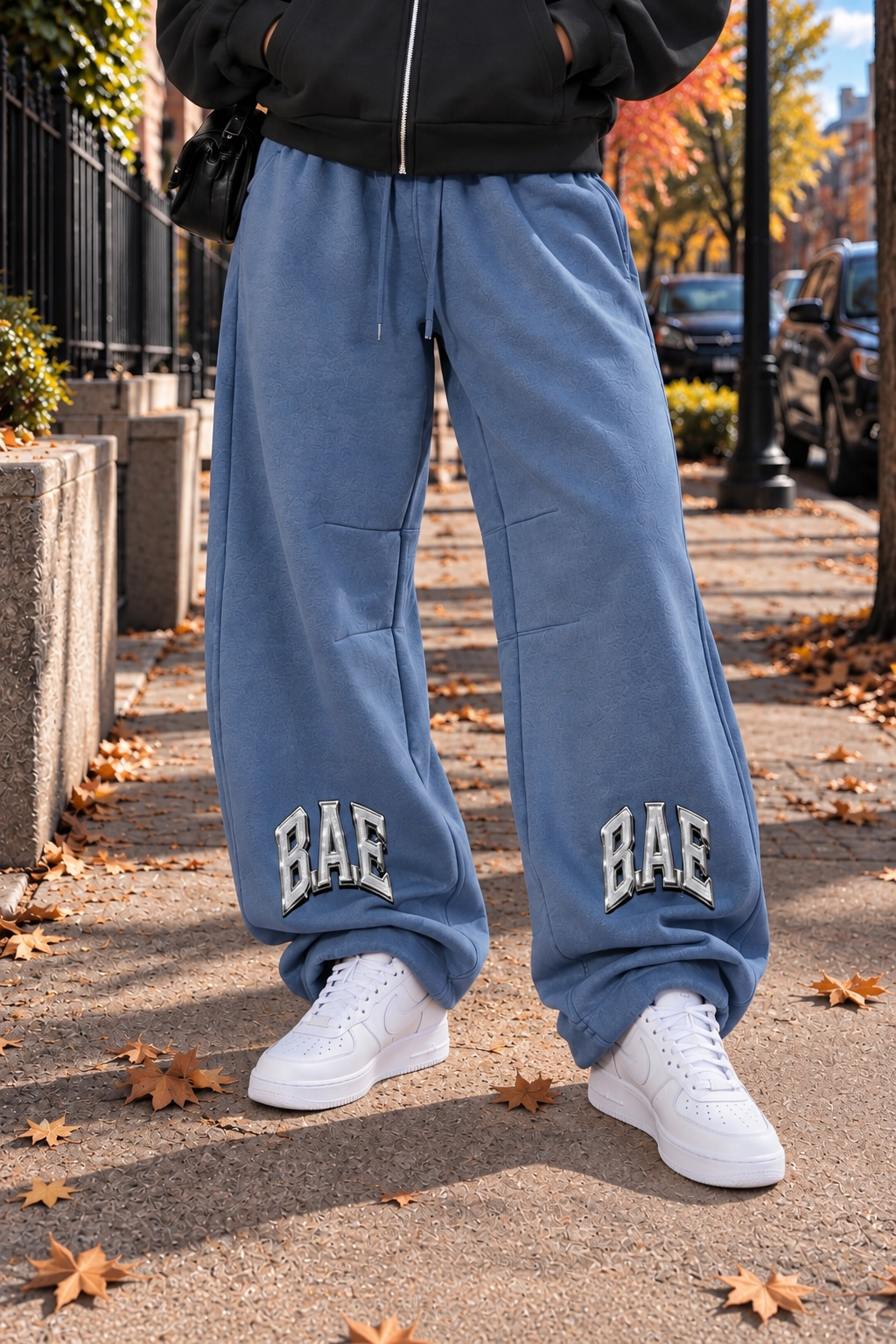 Blue vintage wash B.A.E. Build An Empire sweatpants shown from the waist down, featuring oversized barrel-leg fit, drawstring waist, bold BAE graphics, fleece construction, and crisp white sneakers. Entrepreneur streetwear fashion.