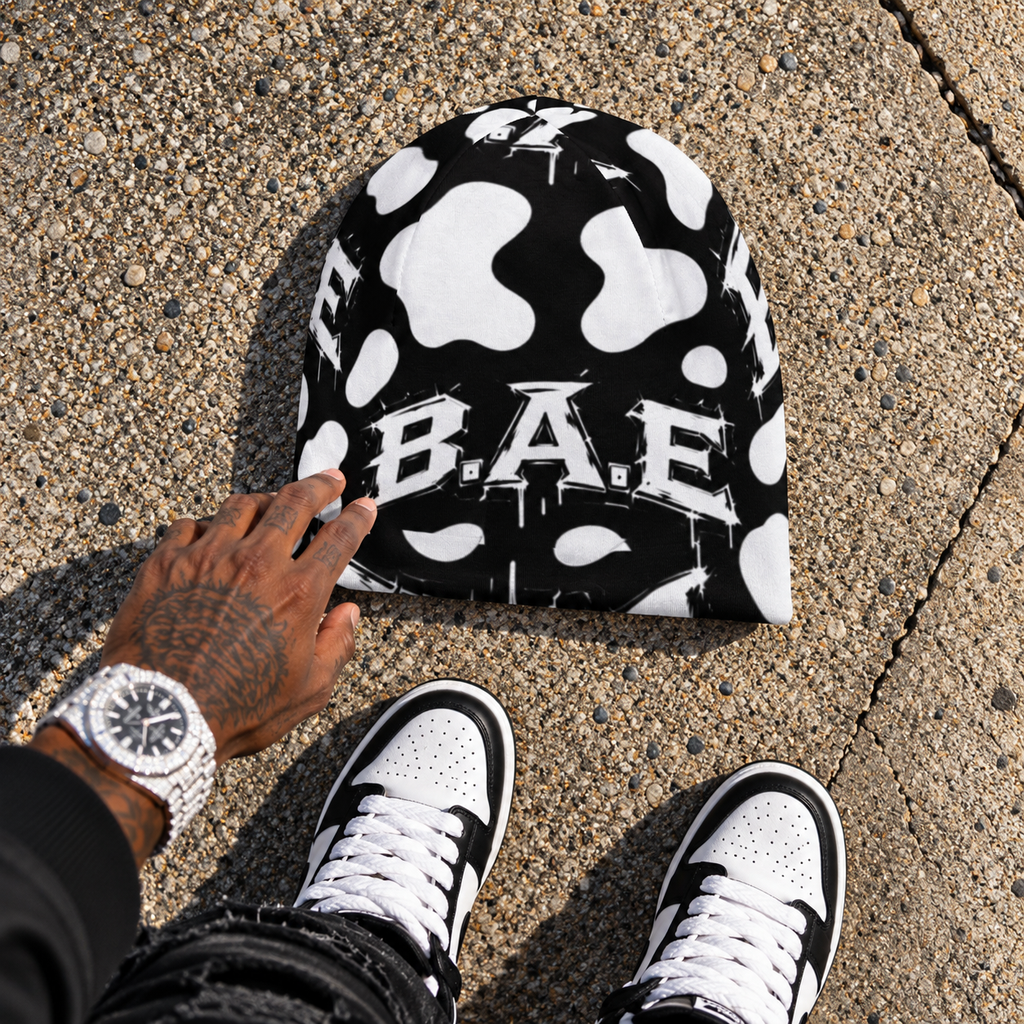 A black beanie with a white cow print pattern and the text 'BAE' across the front, placed on concrete pavement. A tattooed hand is touching the beanie, and the person's feet in black and white sneakers are visible at the bottom of the frame.