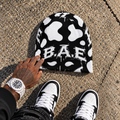 A black beanie with a white cow print pattern and the text 'BAE' across the front, placed on concrete pavement. A tattooed hand is touching the beanie, and the person's feet in black and white sneakers are visible at the bottom of the frame.
