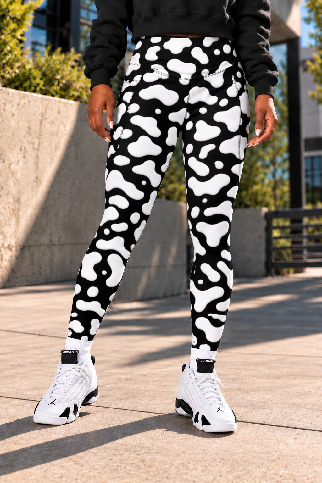 B.A.E. women's black and white cow print leggings with pockets, high-waisted streetwear leggings modeled with a black hoodie and white-and-black sneakers, showing the bold cow-spot pattern, full-length fit, two side pockets, stretchy fabric and statement women's streetwear style.