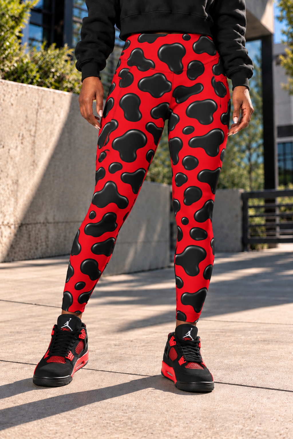 Female model shown from the waist down wearing red and black cow print women's leggings with a black cropped hoodie and black-and-red sneakers, standing on a sunlit concrete walkway and clearly showing the front high-waisted fit, seams, close-fitting silhouette and bold all-over print.