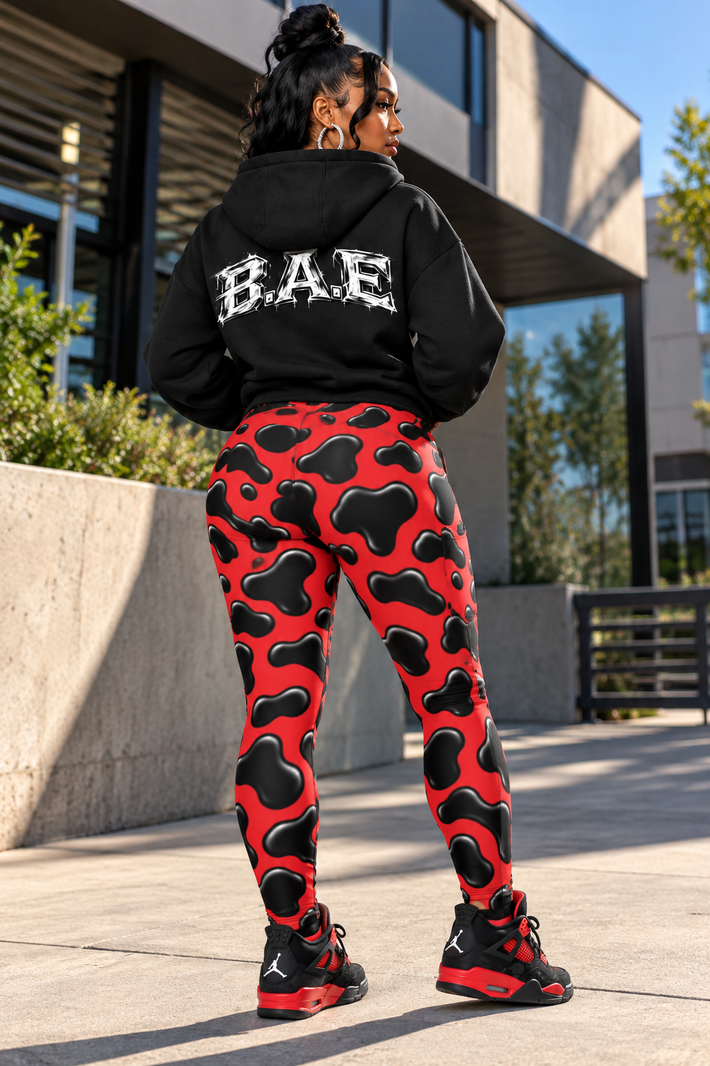Female model viewed from behind wearing red and black cow print women's leggings with a black B.A.E. hoodie and black-and-red sneakers, standing on a sunlit concrete walkway outside modern urban architecture and showing the leggings' full back fit and bold all-over print.