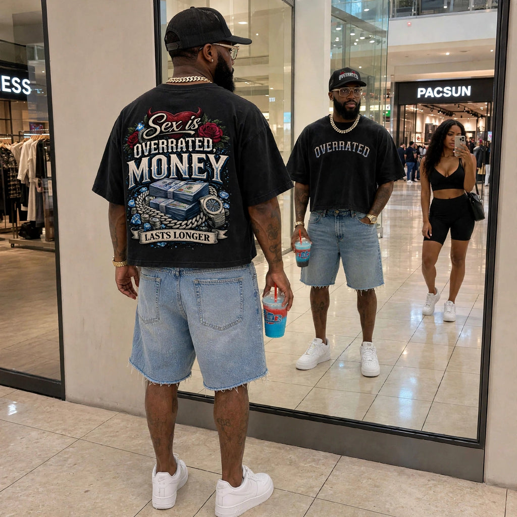 Black oversized boxy crop drop-shoulder t-shirt featuring the “Sex Is Overrated, Money Lasts Longer” back graphic with money, roses, jewelry, and watch artwork. Male model wearing light blue denim shorts, white sneakers, Cuban link chain, luxury watch, and black “OVERRATED” foam trucker hat in a bright shopping mall streetwear lifestyle setting.