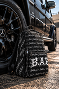 B.A.E. BUILD AN EMPIRE black backpack photographed beside a luxury black Mercedes G-Wagon, showcasing its all-over script pattern, distressed B.A.E. front graphic, textured water-resistant polyester and realistic construction. Premium entrepreneur streetwear backpack for business, college, work and everyday lifestyle.