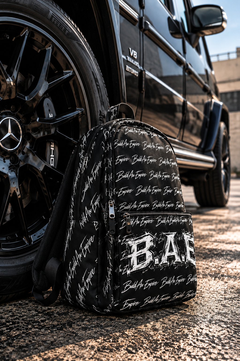 B.A.E. BUILD AN EMPIRE black backpack photographed beside a luxury black Mercedes G-Wagon, showcasing its all-over script pattern, distressed B.A.E. front graphic, textured water-resistant polyester and realistic construction. Premium entrepreneur streetwear backpack for business, college, work and everyday lifestyle.